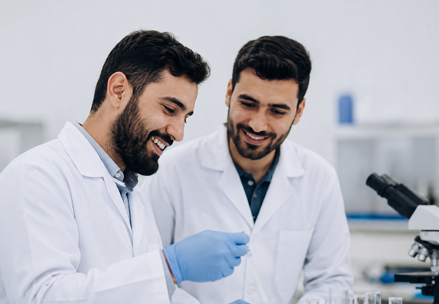 Two chemical laboratory professionals in white lab coats smiling and collaborating at a microscope